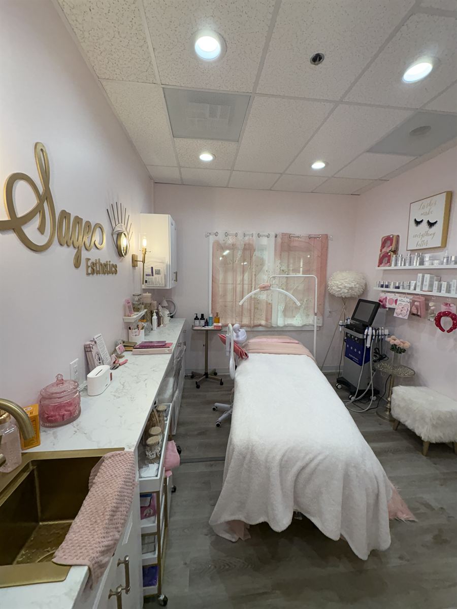 The Sage Esthetics treatment room — the gold Sage sign over the work counter, the treatment bed made up in white and pink, and the product shelves by the window